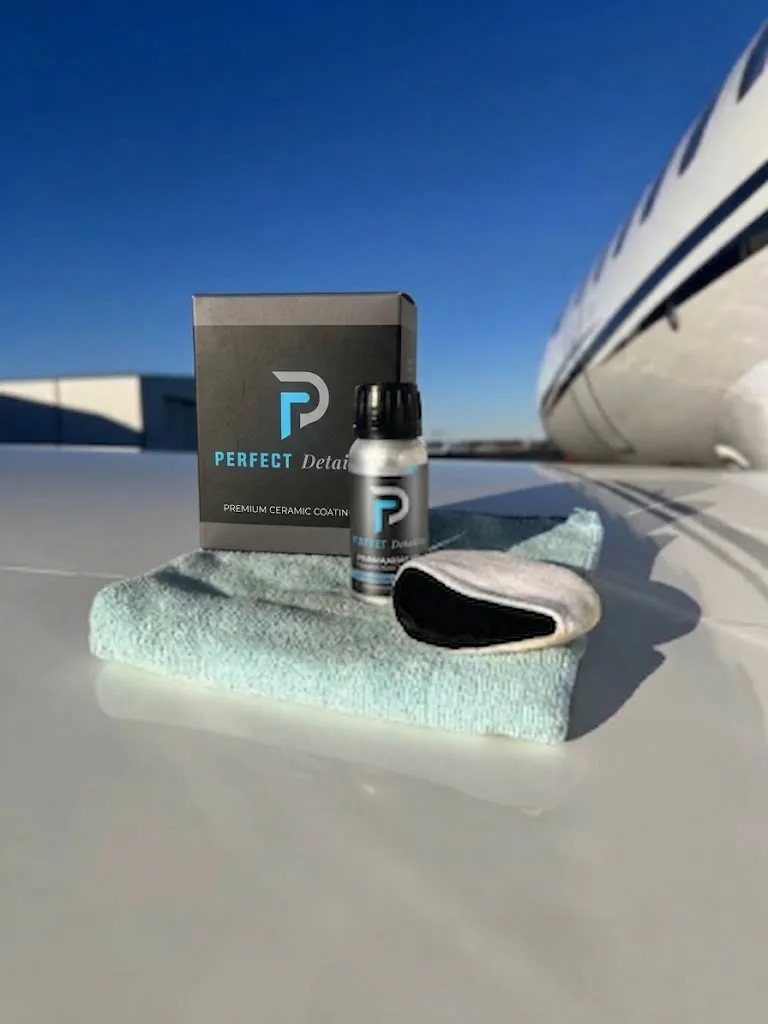 A bottle of Perfect Detailing ceramic coating with its box, a microfibre cloth and an applicator pad, resting on the wing of a business jet under a clear sky