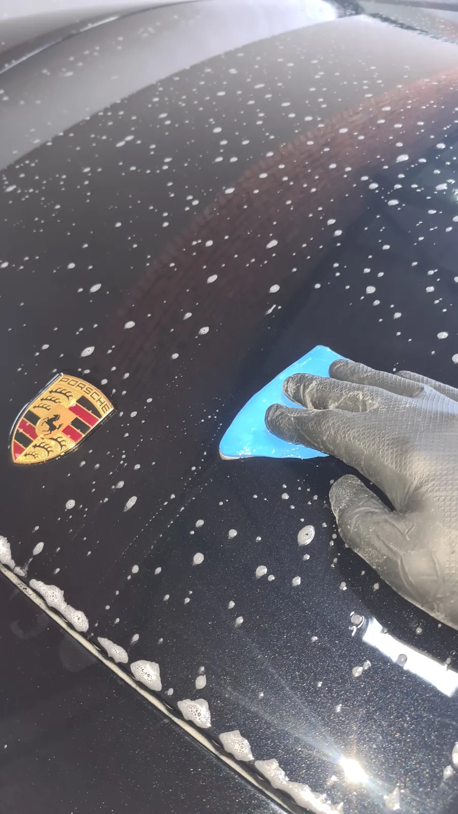 A gloved hand drawing a clay bar across a foamed black panel beside the Porsche crest